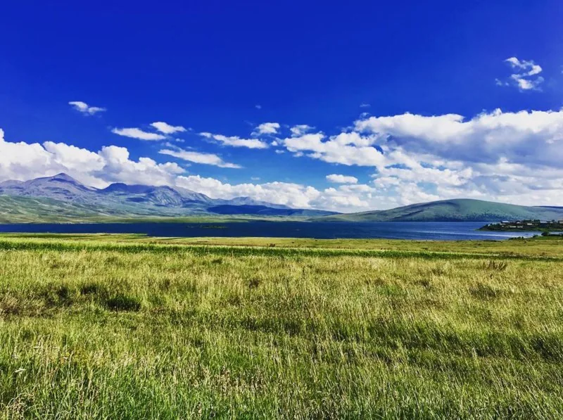Wide green meadow and a lake under a blue sky with cumulus clouds
